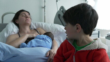 Older sibling touches newborn while mother breastfeeds in hospital bed. Mother is resting, and...