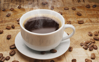 White cup of hot steaming coffee on a wooden table and roasted coffee beans near it.