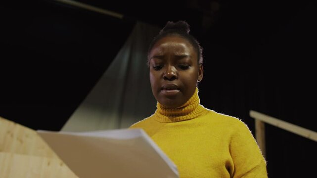 Low angle view of young talented Black actress reading monologue on paper and acting out with expression during rehearsal on theatre stage