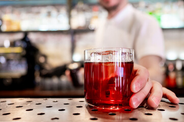 Close-up of a hand holding a glass of red beverage on a bar counter, with an out-of-focus bartender in the background