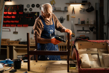 An elderly man in orange gloves and a blue apron sands the leg of a wooden chair with a drill with a special attachment. He is preparing the chair for restoration