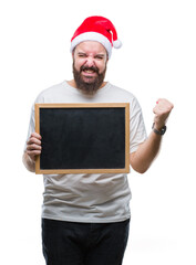 Young caucasian hipster man wearing christmas hat holding blackboard over isolated background screaming proud and celebrating victory and success very excited, cheering emotion