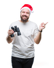 Young caucasian hipster man wearing christmas hat looking though binoculars over isolated...
