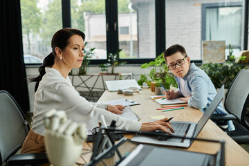 A mother works at her laptop while her son with Down syndrome sits next to her, drawing at a desk in a modern office.