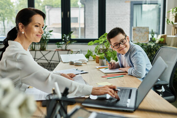 A mother works on her laptop while her son, who has Down syndrome, draws at a nearby table.