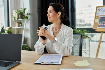 A woman in a white shirt sits at a desk in an office, holding a phone, looking thoughtful.