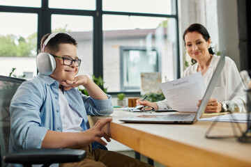 A young boy with Down syndrome works on his laptop alongside his mother in a modern office setting.