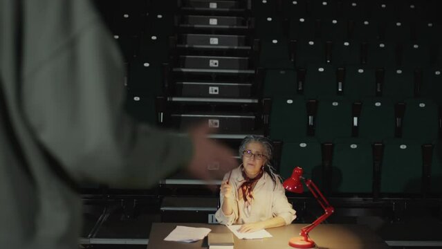 Senior female director sitting in audience in empty theatre and talking with actor on stage during audition