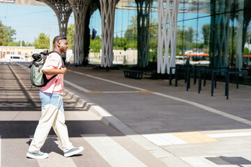 Full height of African man wearing a backpack is walking across a street with modern building of international airport terminal on background.