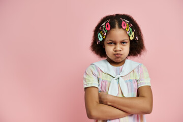 An African American girl with colorful hair clips stands with crossed arms in a pink and white dress on a pink background.
