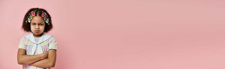 offended African American girl with hair clips stands against a pink backdrop.