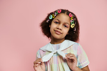 A young African American girl with colorful hair clips smiles while standing on a pink background.