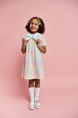 A cute African American girl with hair clips smiles brightly while standing in a rainbow checkered dress against a pink background.