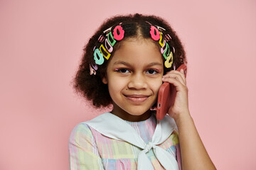 A young African American girl with colorful hair clips stands in a pink dress, smiling while talking on a pink phone.