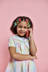 A young African American girl with colorful hair clips stands in a striped dress, smiling and talking on her phone.
