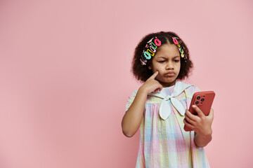 A young African American girl with colorful hair clips is looking intently at her smartphone while standing against a pink background.