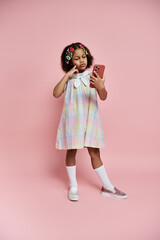 A young girl in a colorful dress stands on a pink background holding a phone.