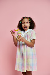 A young African American girl with colorful hair clips stands in a pink dress against a pink backdrop, blowing bubbles with a wand.