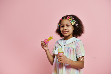 A young African American girl with colorful hair clips blows bubbles in a pink and white dress against a pink background.