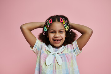 A young African American girl with hair clips stands in a colorful dress against a pink backdrop, radiating joy and happiness.