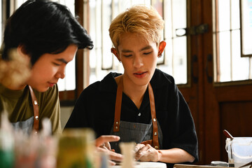 Two friends, one of whom is LGBT, are deeply engrossed in creating art together. They are shaping...