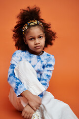 A young African American girl with hair clips in her hair poses in a trendy outfit against an orange background.