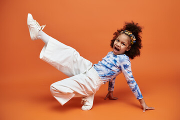 Stylish African American girl with hair clips standing in trendy blue and white shirt and white pants on orange background.