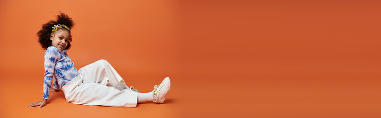 African American girl in a trendy outfit poses against an orange background. She wears white pants, a tie-dye top, and hair clips.