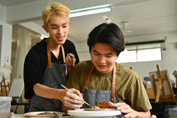 Fototapeta premium Two art students, one sculpting clay on a pottery wheel while the other smiles and offers help, surrounded by art tools and supplies in a creative studio, Crafts and DIY with people concept