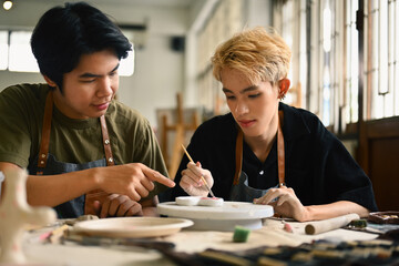Fototapeta premium An art instructor guides an LGBT student as they paint a ceramic piece. Both are focused in a bright studio filled with pottery tools and materials, fostering a supportive learning environment