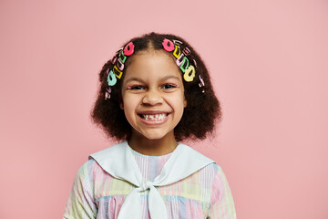 A young African American girl with colorful hair clips smiles brightly while standing in a pink dress against a pink background.