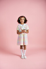 A young African American girl in a pastel dress with hair clips stands on a pink background.