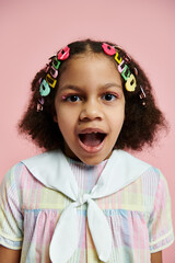 A young African American girl with colorful hair clips stands in a pink dress with a surprised expression on her face.