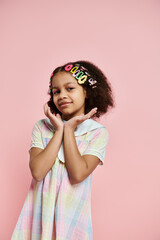 A cute African American girl with hair clips stands in a colorful dress against a pink background.