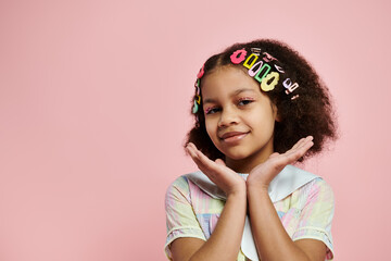 Young Black girl with cute hairstyle and colorful hair clips poses in pastel dress on pink background.