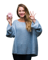 Obraz premium Young beautiful blonde woman eating sweet donut over isolated background doing ok sign with fingers, excellent symbol