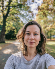 Natural Looking 30 Year Old Woman Taking a Mobile Photo Selfie Outdoors with Casual Clothes, Relaxed Expression and Pose, Surrounded by Trees in Sunlit Park