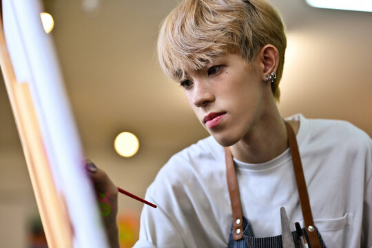 A focused LGBT artist with blonde hair carefully works on a painting with a brush. The close-up captures the intensity and precision of the artist in a brightly lit studio