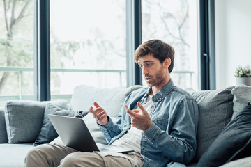 Concentrated 30s man sitting on sofa in living room looking at computer screen, involved in online video call working issues negotiations with colleagues or giving professional consultation to client