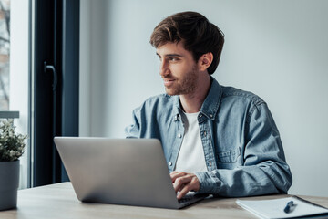 Side view handsome young businessman working with computer remotely, sitting at wooden table in office. Pleasant happy man communicating in social network, searching information online..