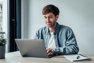 Side view handsome young businessman working with computer remotely, sitting at wooden table in office. Pleasant happy man communicating in social network, searching information online..