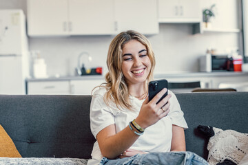 Happy pleasant millennial girl relaxing on comfortable couch, holding smartphone in hands. Smiling young lady chatting in social networks, watching funny videos, using mobile applications at home..