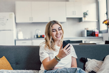 Happy pleasant millennial girl relaxing on comfortable couch, holding smartphone in hands. Smiling young lady chatting in social networks, watching funny videos, using mobile applications at home..