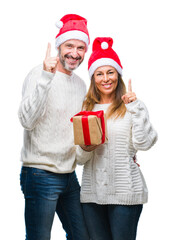 Middle age hispanic couple wearing christmas hat and holding gift over isolated background surprised with an idea or question pointing finger with happy face, number one