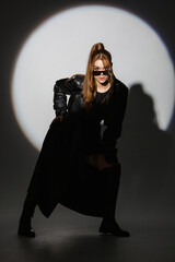 model girl in a black short dress in leather raincoat with glasses in studio with hard spotlight...