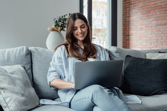 Concentrated millennial girl sit on couch working on laptop browsing internet at home during lazy weekend, focused young woman freelancer busy using computer surfing wireless web shopping online.