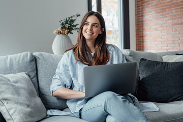 Concentrated millennial girl sit on couch working on laptop browsing internet at home during lazy...