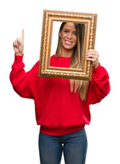 Fototapeta premium Beautiful young woman holding vintage frame surprised with an idea or question pointing finger with happy face, number one