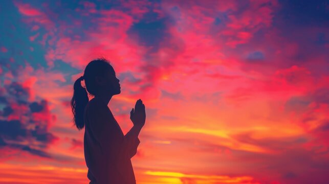 A silhouette of a woman praying at sunset with the sky turned orange and pink, perfect for inspirational or faith-related themes