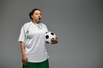 A female soccer referee holding a ball, with a whistle in her mouth, is ready to start the game.